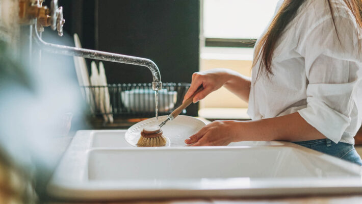 Junge Frau wäscht Geschirr mit Kalk mit Holzbürste mit natürlichen Borsten am Fenster in der Küche