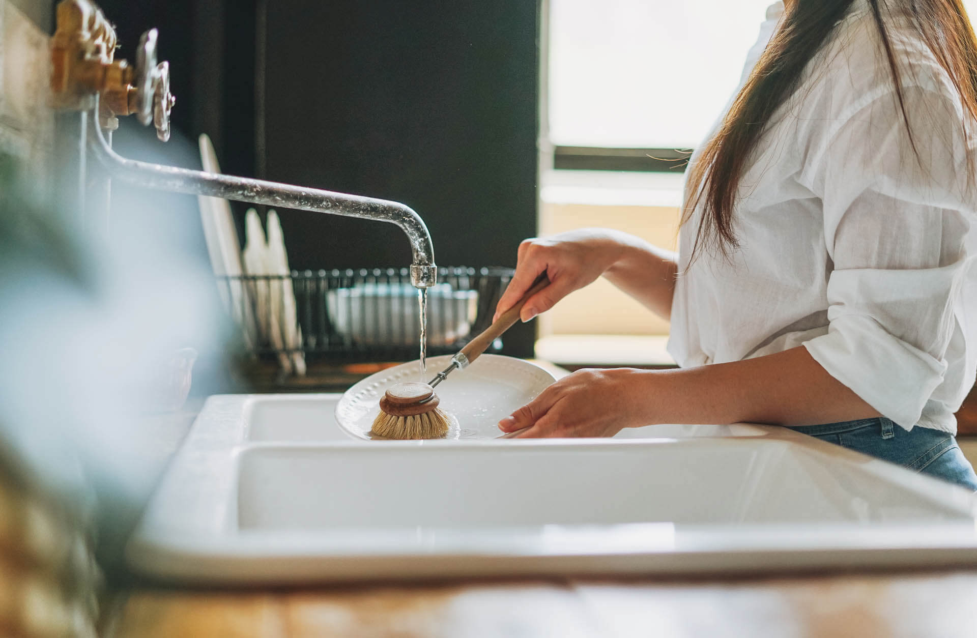 Junge Frau wäscht Geschirr mit Kalk mit Holzbürste mit natürlichen Borsten am Fenster in der Küche